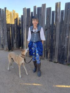 photo of Kari Carlisle in Steampunk outfit, smailing at the camera and holding her leashed fawn colored greyhound, Myka, who is looking at her expectantly.